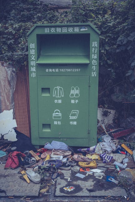 A green, old-style clothing collection bin situated outdoors, with Chinese characters and a contact phone number on the front panel. The bin features separate compartments labeled for items such as cl